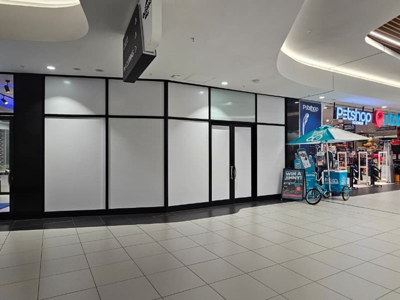A vacant retail storefront unit located within a modern shopping mall, featuring large glass panels and high-quality tiled flooring.