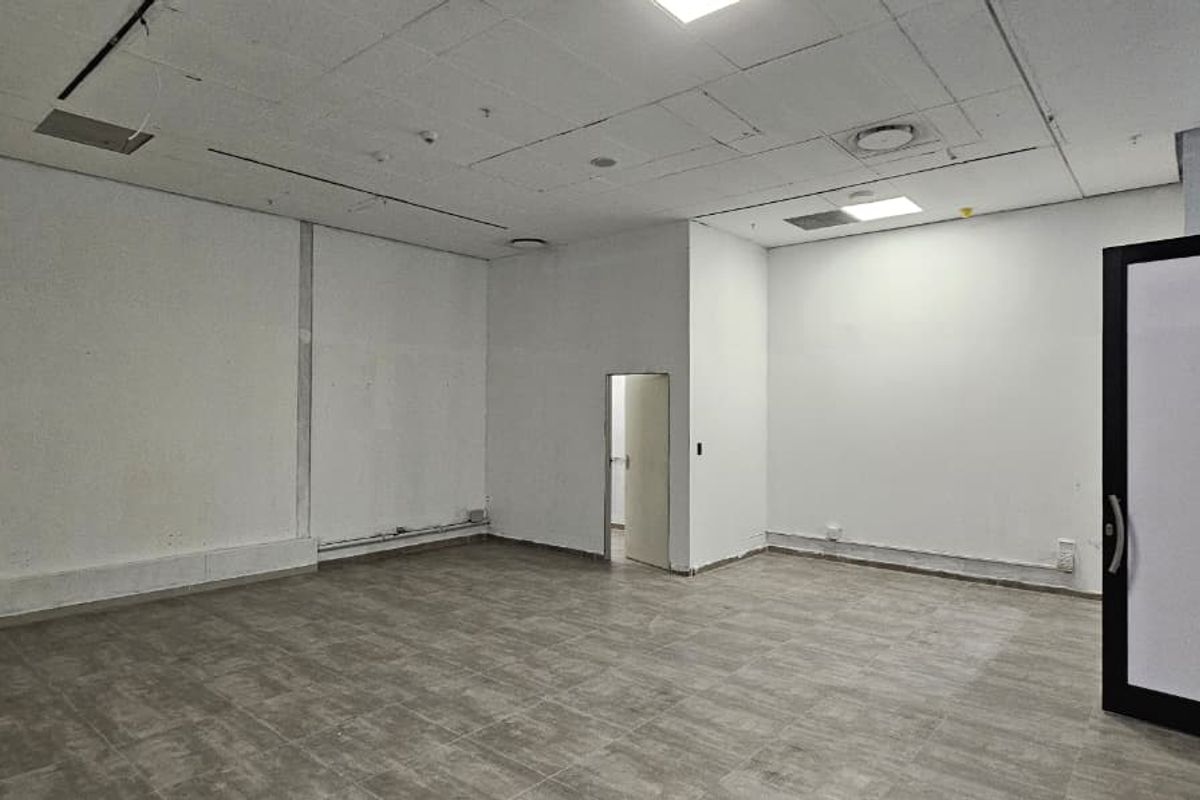 An empty, open-plan retail space featuring tiled flooring, a suspended ceiling with recessed lighting, and a glass entrance door.