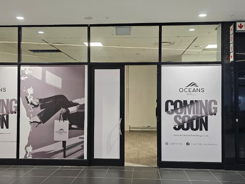 A vacant retail storefront unit featuring large glass display windows, tiled flooring, and recessed ceiling lighting, located within a shopping mall.