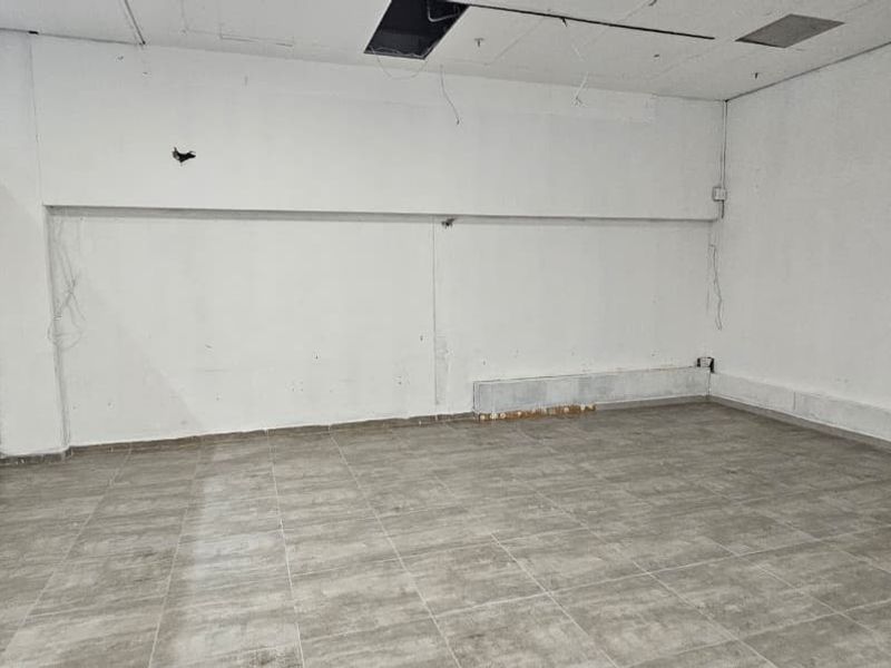 An empty, open-plan retail space featuring tiled flooring and a large storefront window, currently requiring ceiling and wall repairs.