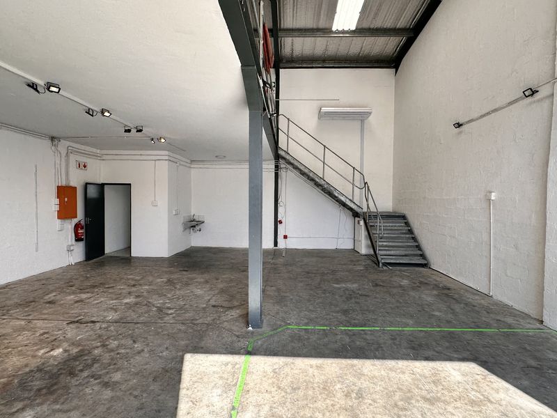 Spacious industrial open-plan unit featuring concrete flooring, exposed ceiling structure, a steel staircase, and a small kitchenette area.