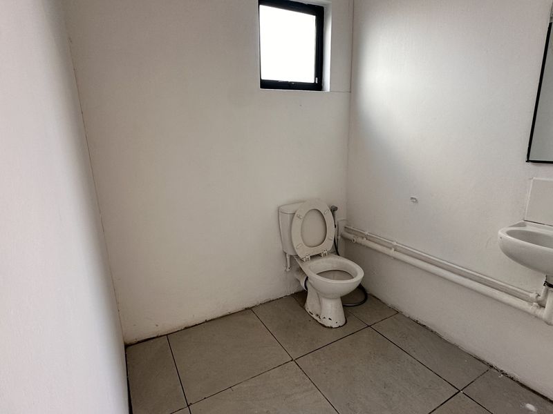 A functional, minimalist bathroom featuring a toilet, wall-mounted basin, and tiled flooring, currently in need of cosmetic updates and finish improvements.