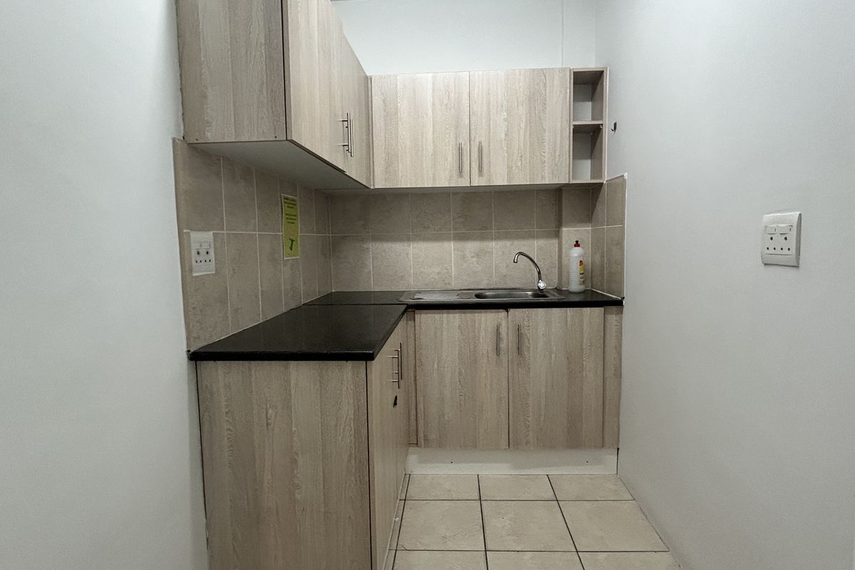 A compact kitchenette featuring light wood-grain cabinetry, dark laminate countertops, a stainless steel sink, and neutral tiled flooring and backsplash.