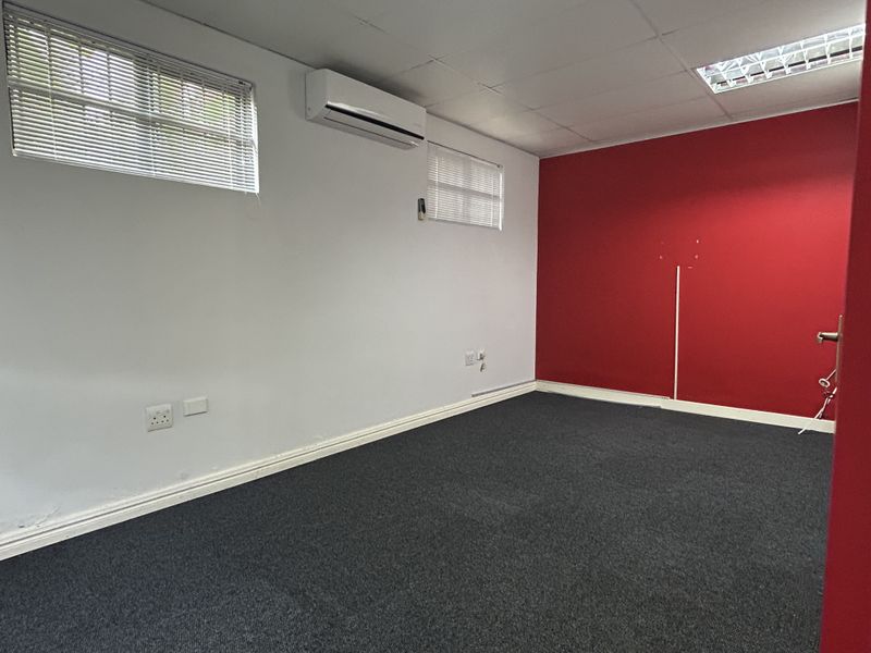 A vacant office space featuring dark carpeting, a split-system air conditioner, drop ceiling with integrated lighting, and a bold red accent wall.