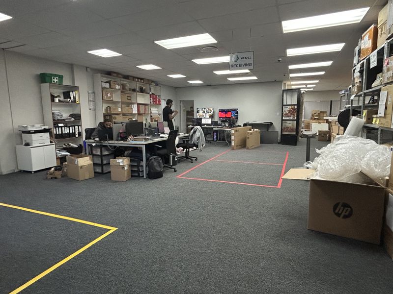 A functional commercial office space featuring suspended ceilings, recessed lighting, and carpeted flooring, currently utilized as a receiving and administrative work area.