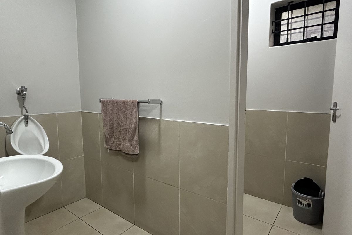 A clean, functional commercial bathroom featuring a pedestal sink, wall-mounted toilet, and neutral tiled finishes with a secure window.