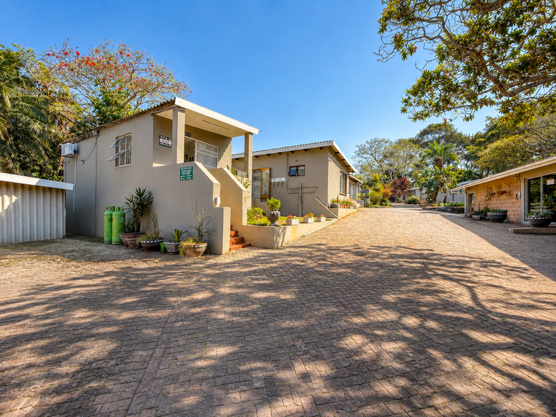 A well-maintained lodge exterior featuring a spacious paved driveway, covered entrance areas, and established garden surroundings under a clear blue sky.