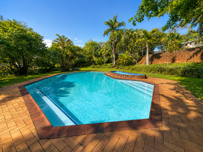 A well-maintained swimming pool area featuring brick paving, a raised pool edge, and lush surrounding garden landscaping.