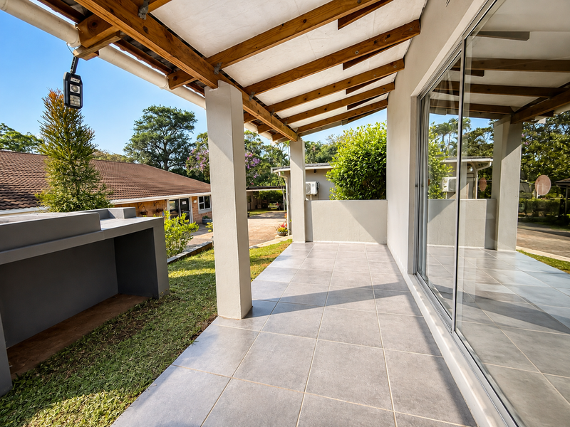 A covered outdoor patio featuring tiled flooring, a built-in braai, and a timber pergola roof, providing a seamless transition to the garden area.