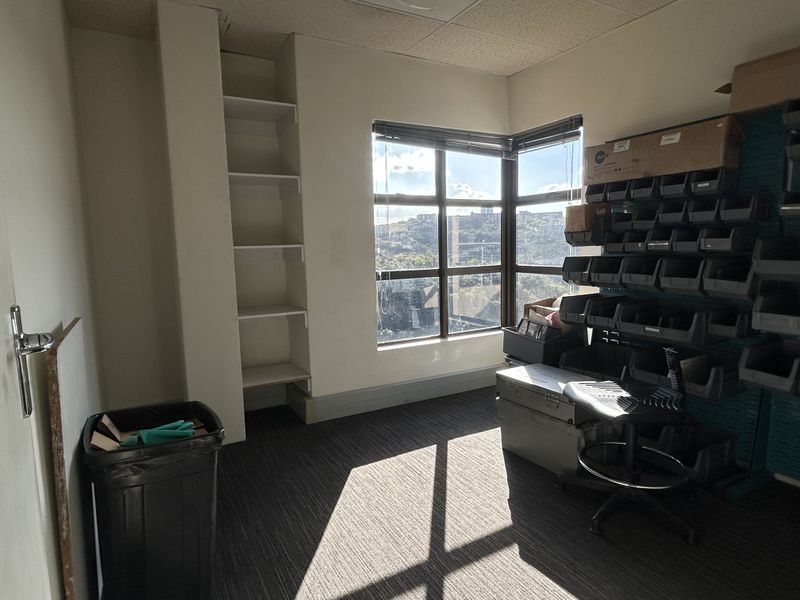 A functional office space featuring built-in shelving, a corner window with natural light, and carpeted flooring, suitable for a professional workspace.