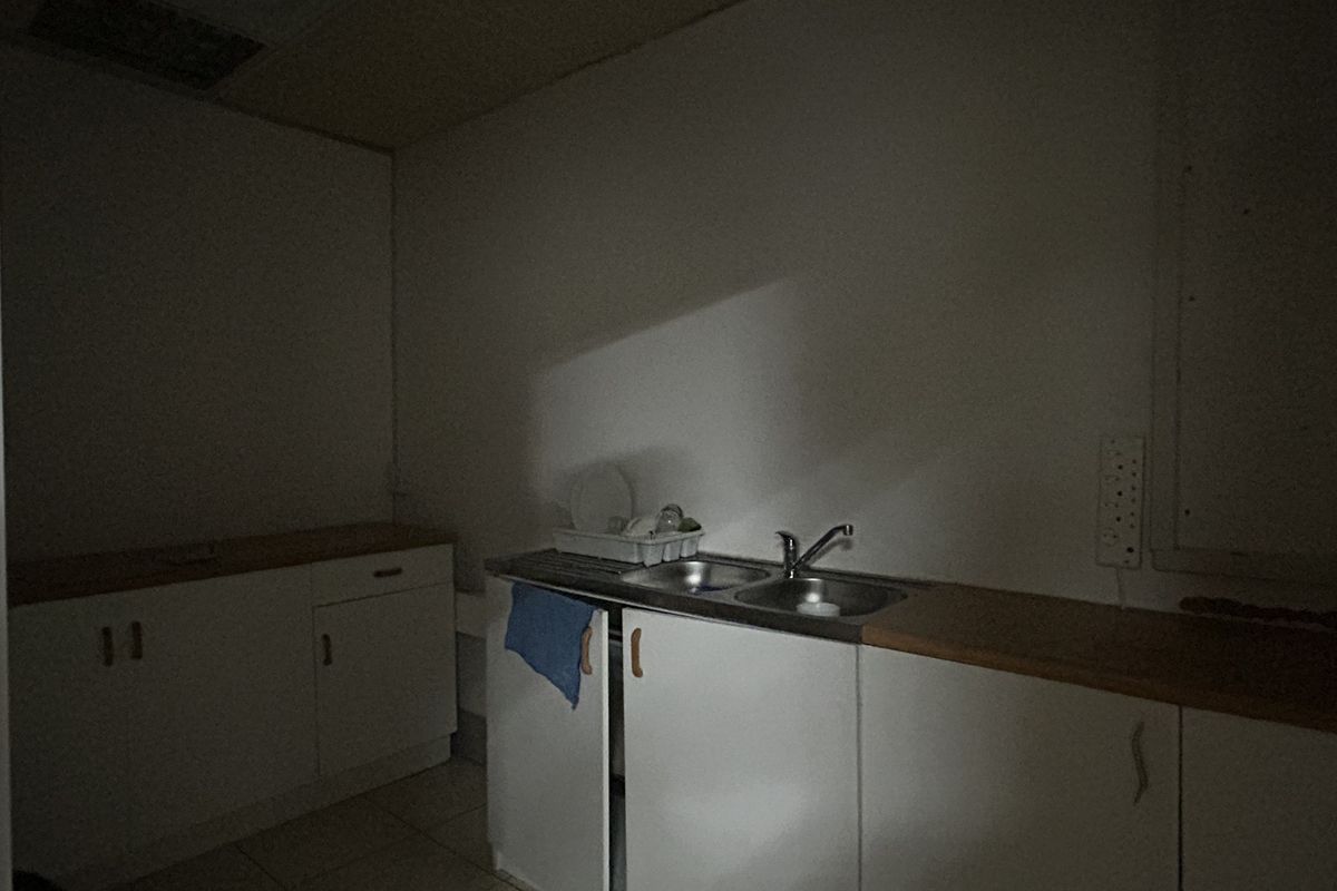 A small office kitchenette featuring basic built-in cabinetry, laminate countertops, and a stainless steel sink, requiring improved lighting and cosmetic updates.