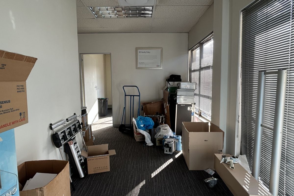 A cluttered office space featuring carpeted flooring, suspended ceiling tiles, and large windows with venetian blinds, currently used for storage.