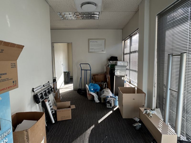 A cluttered office space featuring carpeted flooring, suspended ceiling tiles, and large windows with venetian blinds, currently used for storage.
