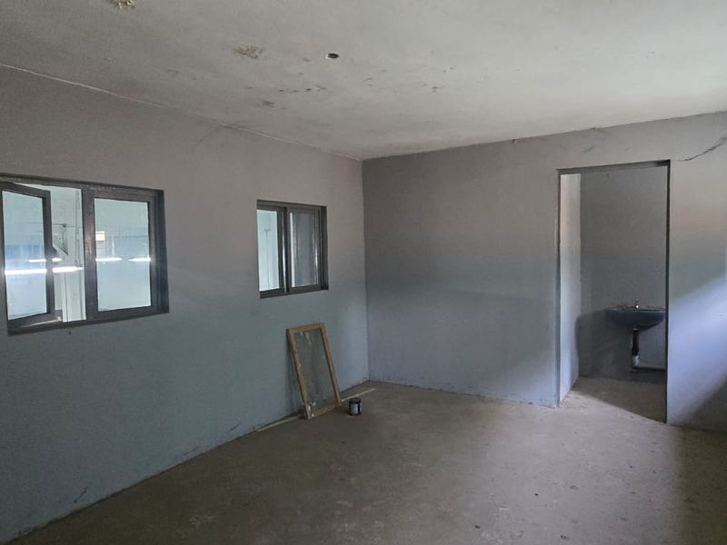 An empty, unfinished utility space featuring grey painted walls, aluminium window frames with security bars, and a small wall-mounted basin in the adjacent alcove.