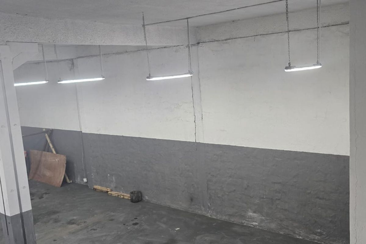 An open-plan industrial utility space featuring concrete flooring, exposed fluorescent lighting, and structural support columns, suitable for storage or workshop use.
