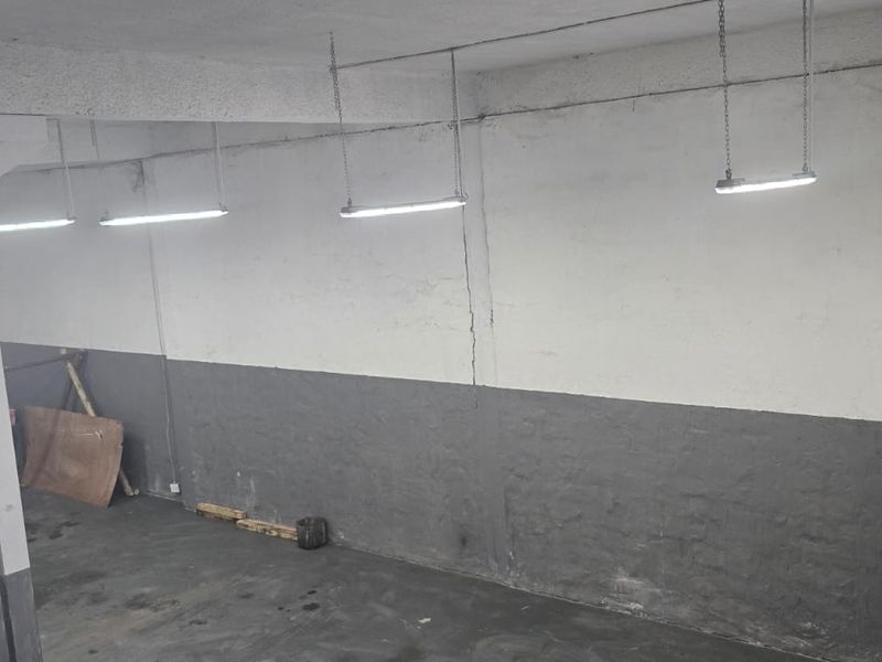 An open-plan industrial utility space featuring concrete flooring, exposed fluorescent lighting, and structural support columns, suitable for storage or workshop use.