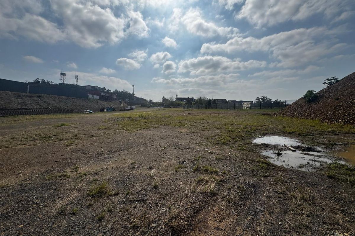 A wide-angle view of a vacant commercial land plot featuring a retaining wall, perimeter fencing, and uneven terrain with patches of standing water.
