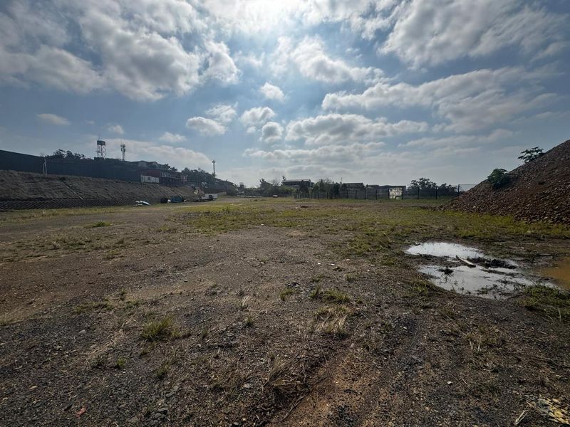 A wide-angle view of a vacant commercial land plot featuring a retaining wall, perimeter fencing, and uneven terrain with patches of standing water.