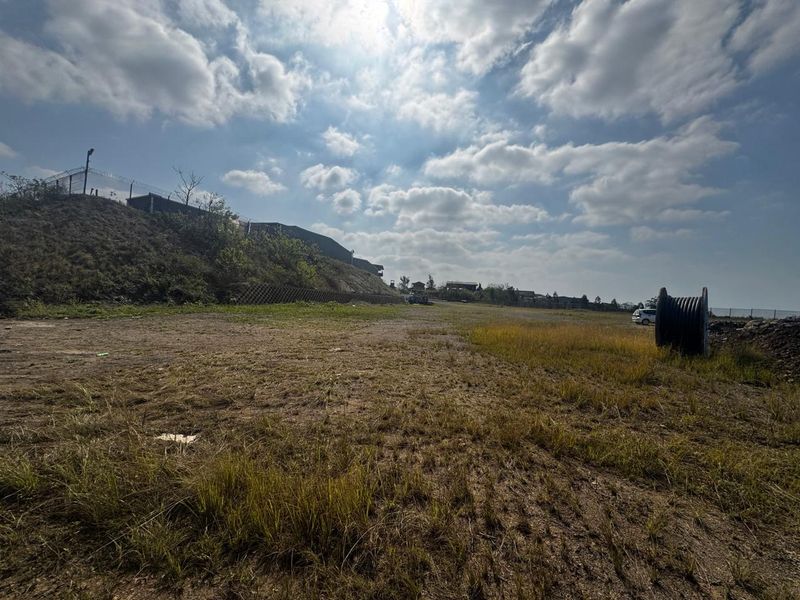 A wide-angle view of a vacant commercial land plot featuring a retaining wall, perimeter security fencing, and open grass terrain.