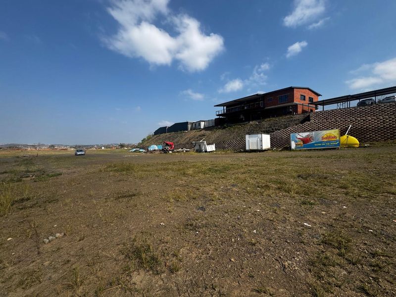 A wide-angle view of a vacant commercial land plot featuring an elevated building structure, a large retaining wall, and an existing billboard.