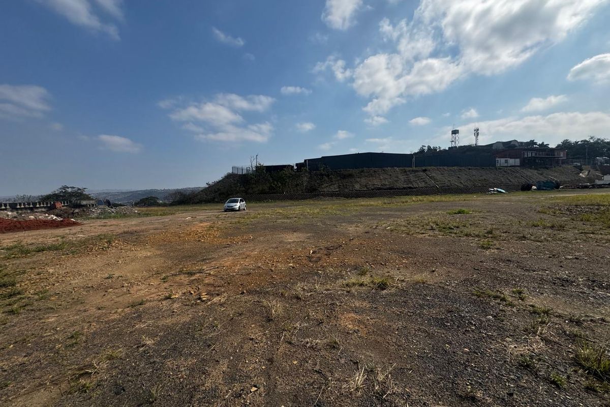 A wide-angle view of a vacant commercial land plot featuring a retaining wall and perimeter fencing under a clear sky.