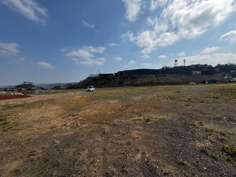 A wide-angle view of a vacant commercial land plot featuring a retaining wall and perimeter fencing under a clear sky.