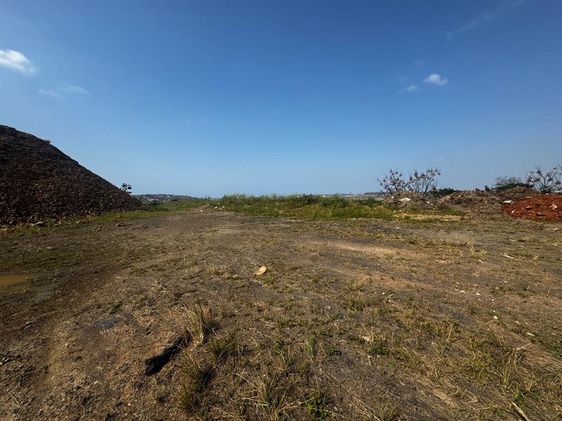 A wide-angle view of a vacant commercial land plot featuring cleared earth, uneven terrain, and surrounding mounds of soil under a clear sky.