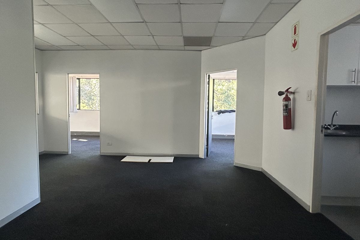 An empty office space featuring a suspended ceiling, dark carpeting, and access to a small kitchenette area.