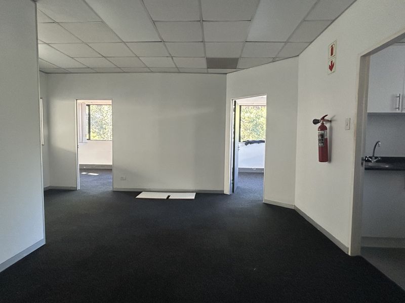 An empty office space featuring a suspended ceiling, dark carpeting, and access to a small kitchenette area.