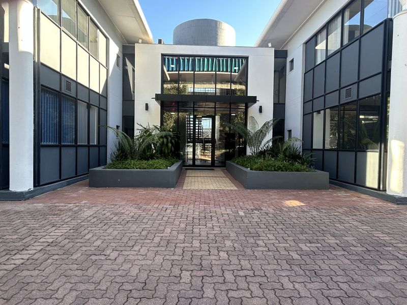 Modern office building entrance featuring a glass facade, paved driveway, and integrated landscaping planters.