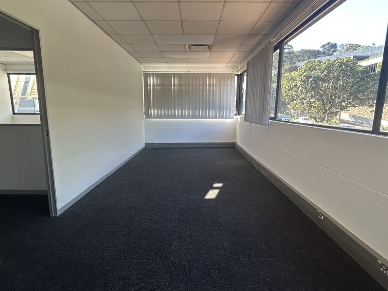 A bright, empty office space featuring carpeted flooring, a suspended ceiling, and large windows with vertical blinds, offering a clean professional environment.