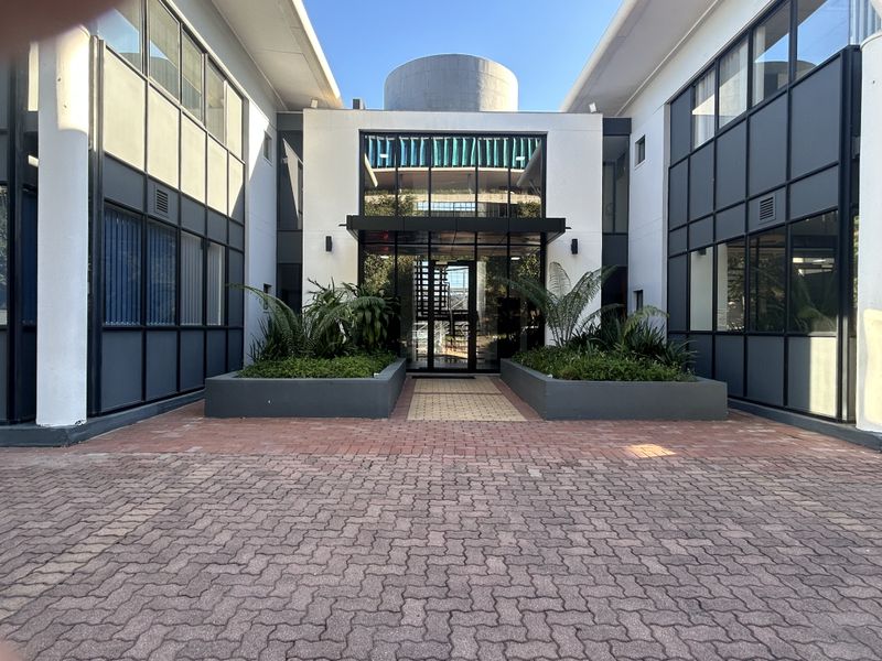 Modern office building entrance featuring a glass facade, paved driveway, and integrated landscaping planters.