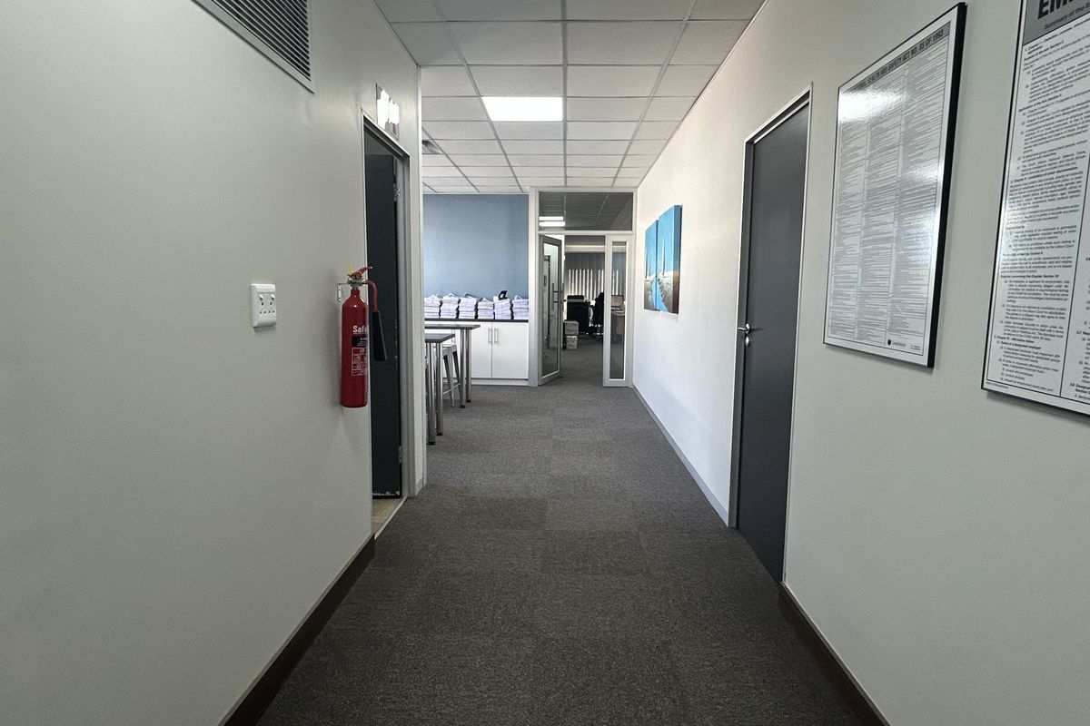 A clean, well-lit office hallway featuring carpeted flooring, a suspended ceiling with recessed lighting, and glass partition doors leading to adjacent workspaces.