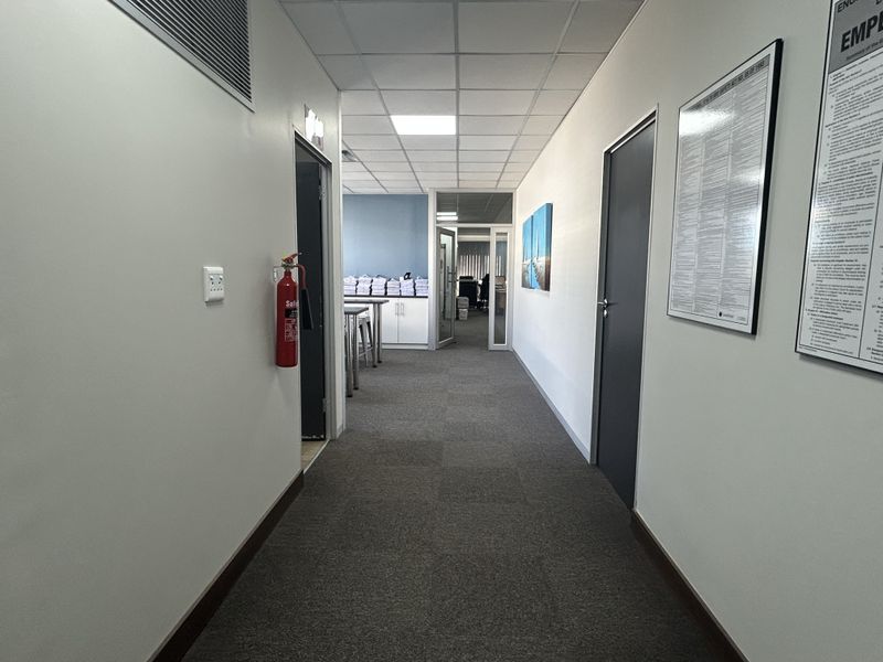 A clean, well-lit office hallway featuring carpeted flooring, a suspended ceiling with recessed lighting, and glass partition doors leading to adjacent workspaces.