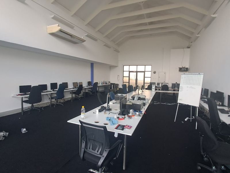 Spacious open-plan office area featuring high vaulted ceilings with exposed rafters, wall-mounted air conditioning, and large windows providing natural light.