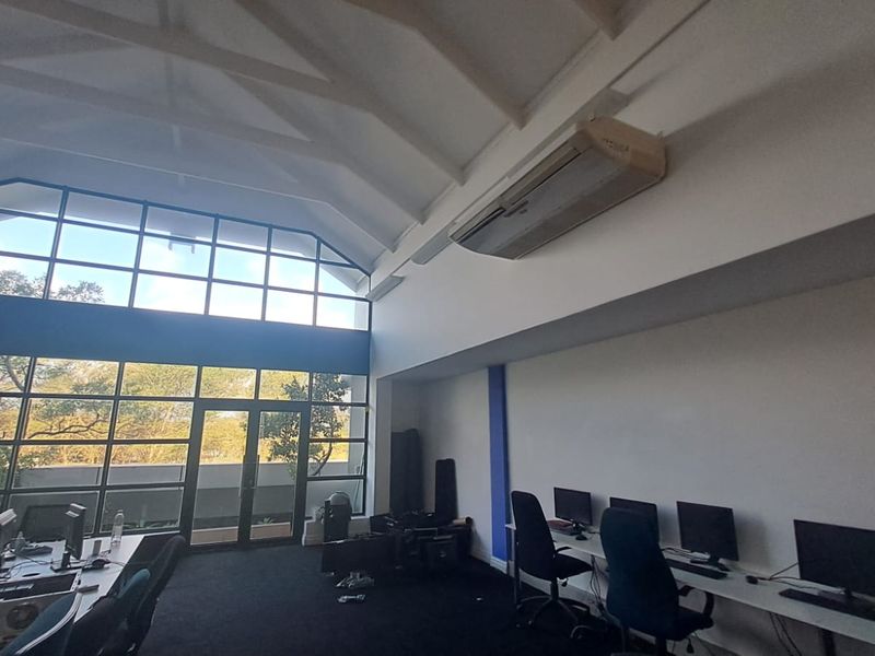 Spacious office area featuring a high vaulted ceiling, large glass windows providing natural light, and a wall-mounted air conditioning unit.