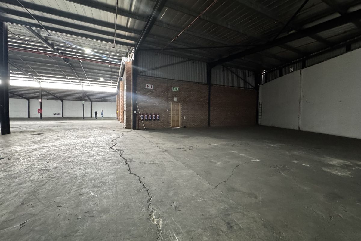 Expansive industrial warehouse space featuring high ceilings, exposed steel structure, brick walls, and concrete flooring with visible surface cracking.