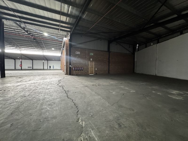 Expansive industrial warehouse space featuring high ceilings, exposed steel structure, brick walls, and concrete flooring with visible surface cracking.