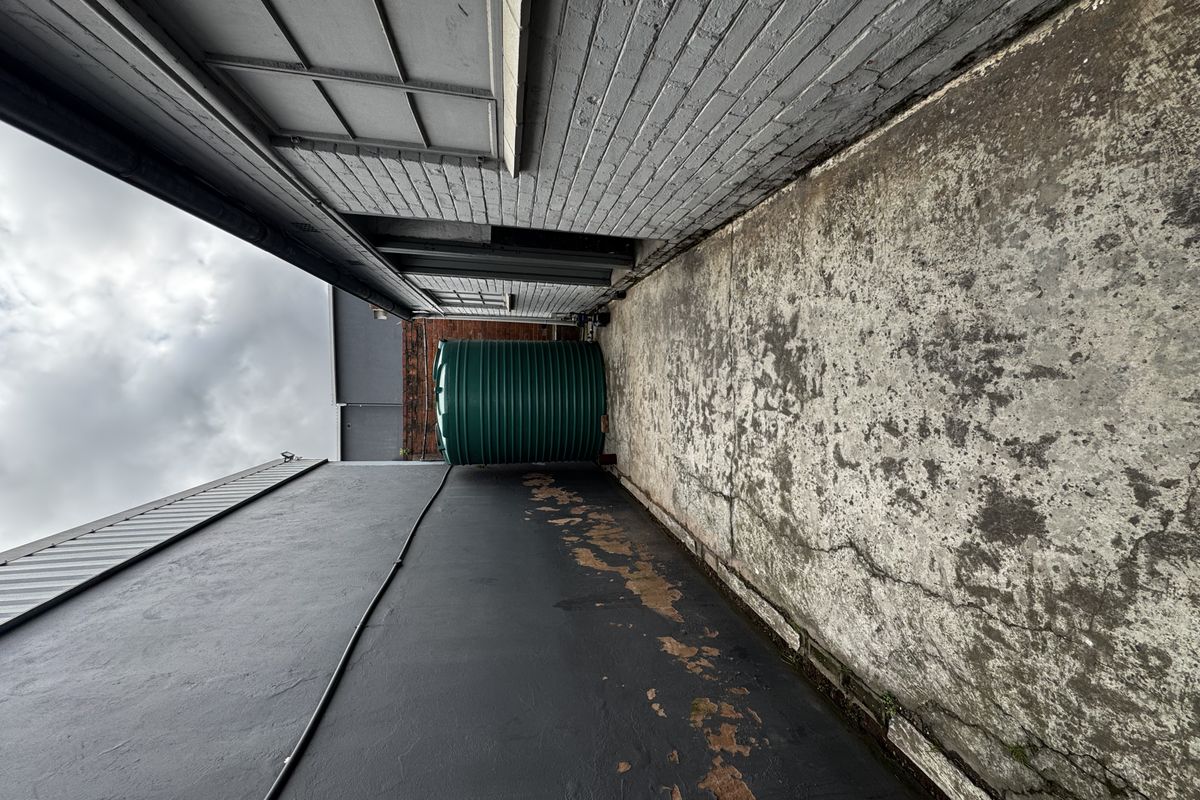 A narrow exterior side passage featuring a water storage tank, concrete paving, and painted brick walls showing signs of wear.