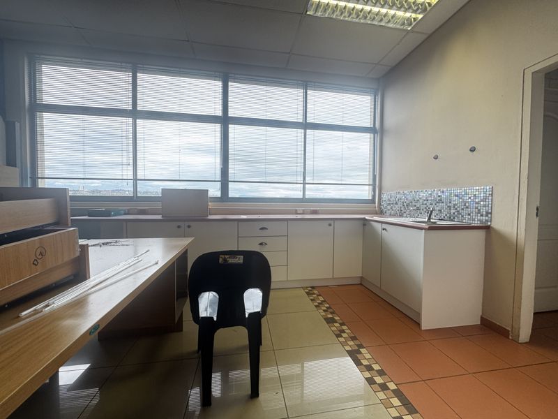 A functional office kitchenette featuring built-in white cabinetry, a stainless steel sink, mosaic backsplash, and large windows with venetian blinds.
