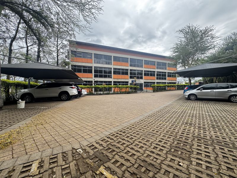 A multi-story office building exterior featuring a paved parking area with covered carports and mature landscaping.