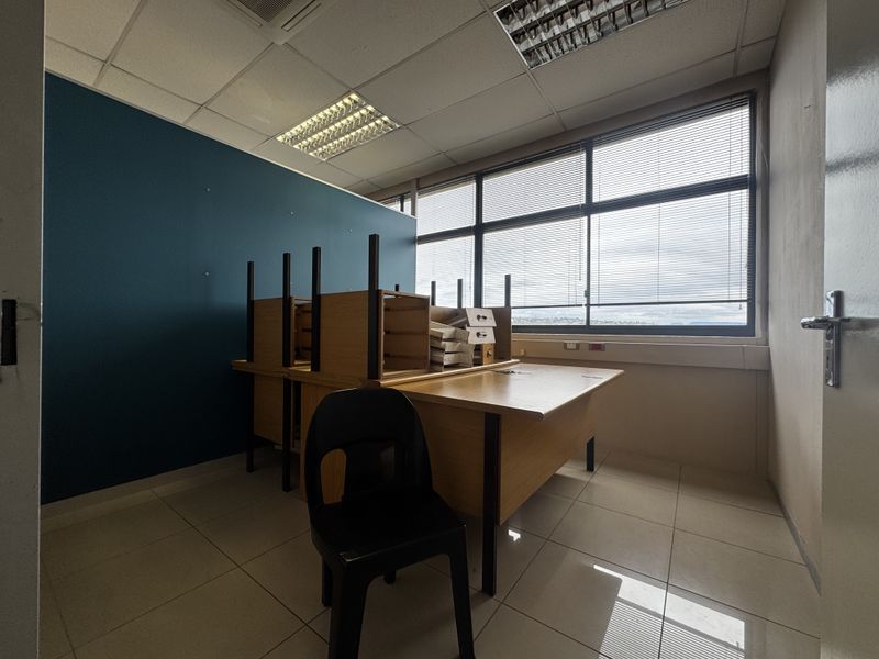 A functional office space featuring tiled flooring, a large window with blinds, and recessed ceiling lighting, providing a bright and professional work environment.