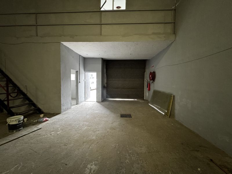 Industrial warehouse space featuring a roller shutter door, mezzanine level with metal staircase, and concrete flooring, suitable for storage or workshop use.