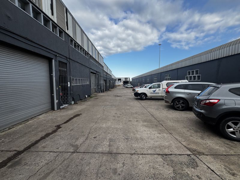 Exterior view of an industrial complex featuring a concrete driveway, multiple roller shutter doors, and secure access points.