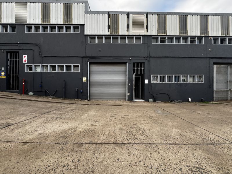 Industrial warehouse exterior featuring a large roller shutter door, security access points, and durable metal cladding with a concrete driveway.