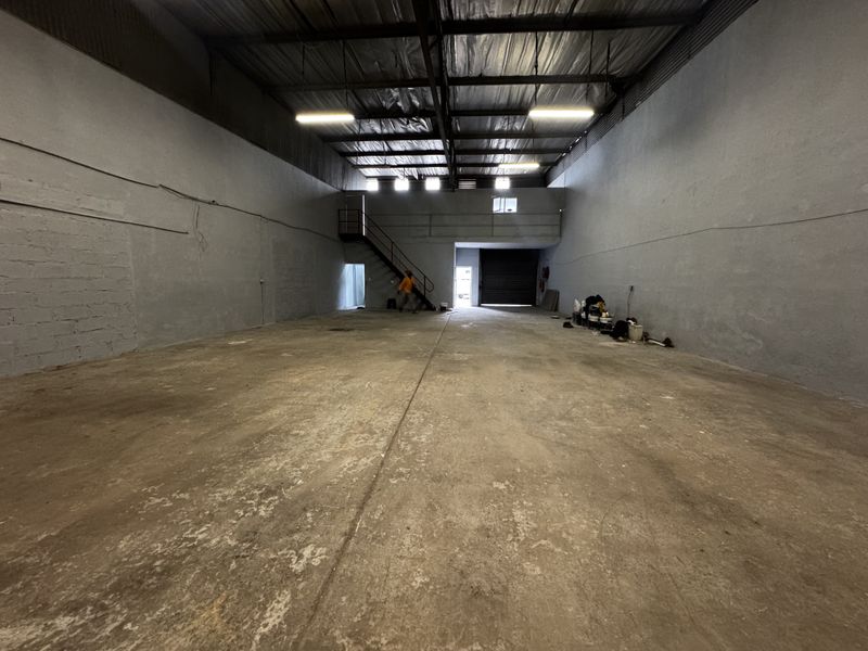 Spacious industrial warehouse unit featuring high ceilings, concrete flooring, a mezzanine level with staircase access, and a large roller shutter door for loading.