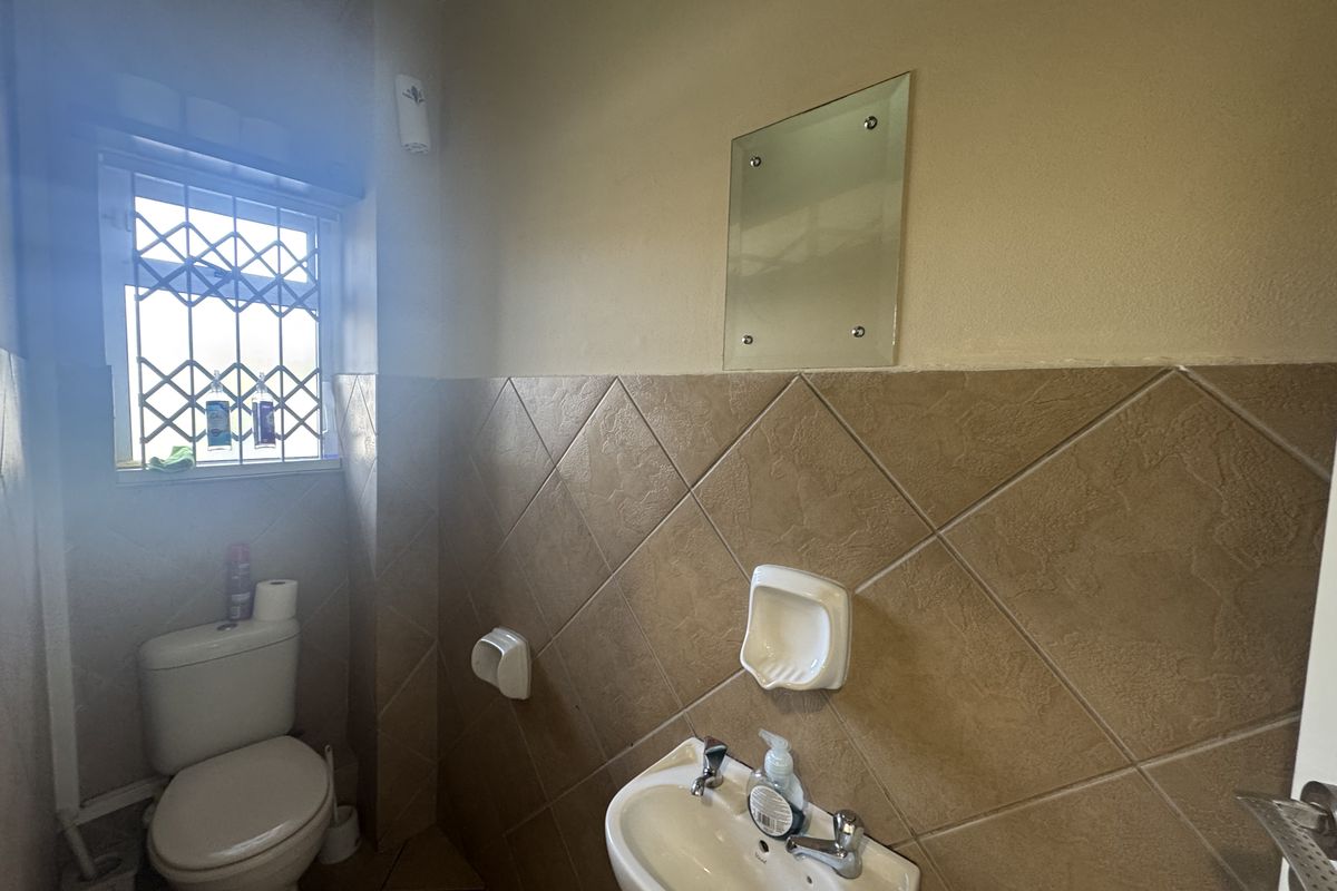 A functional office bathroom featuring a toilet, wall-mounted washbasin, tiled walls, and a security gate on the window.