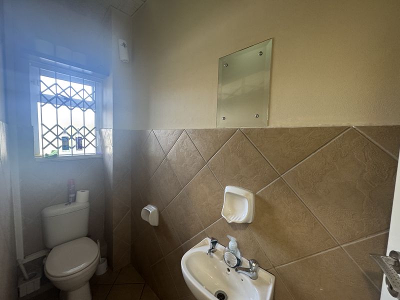 A functional office bathroom featuring a toilet, wall-mounted washbasin, tiled walls, and a security gate on the window.