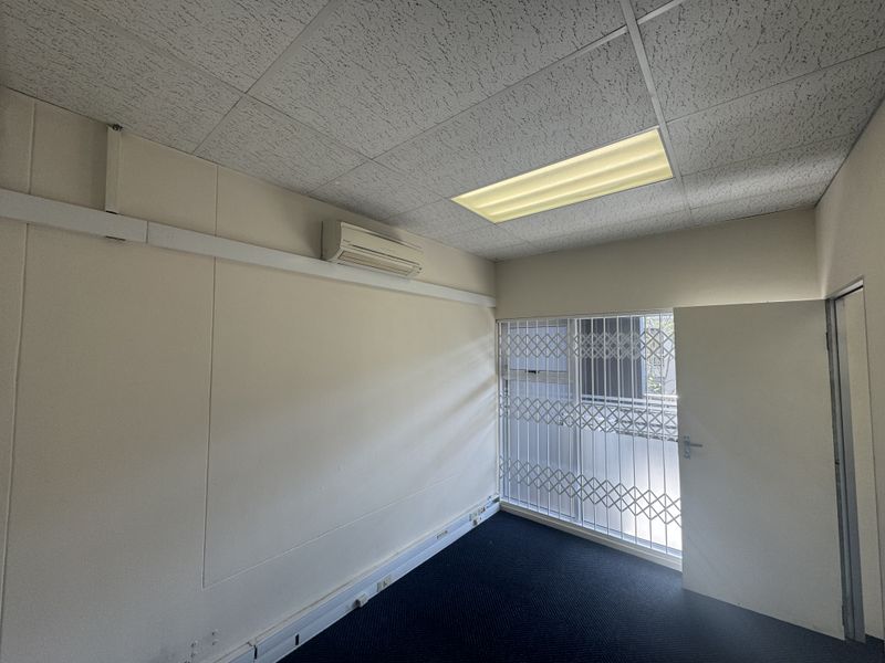 A vacant office space featuring a suspended ceiling, recessed lighting, wall-mounted air conditioning, and a security gate over the window.