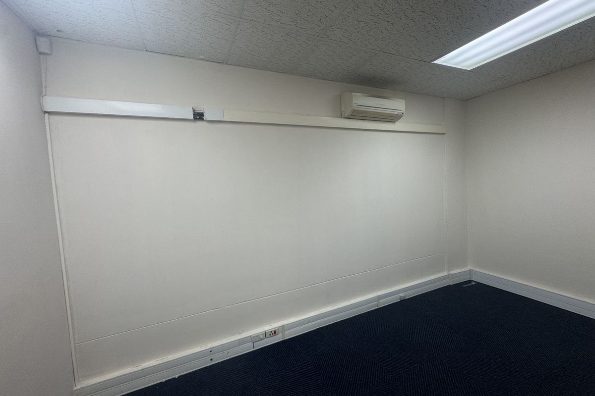 An empty office space featuring a suspended ceiling, recessed lighting, wall-mounted air conditioning, and blue carpeted flooring.